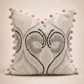 A square decorative throw pillow in a light beige fabric with a textured damask pattern, featuring black embroidered heart motifs and a border of pink pom-poms, set against a solid beige background.