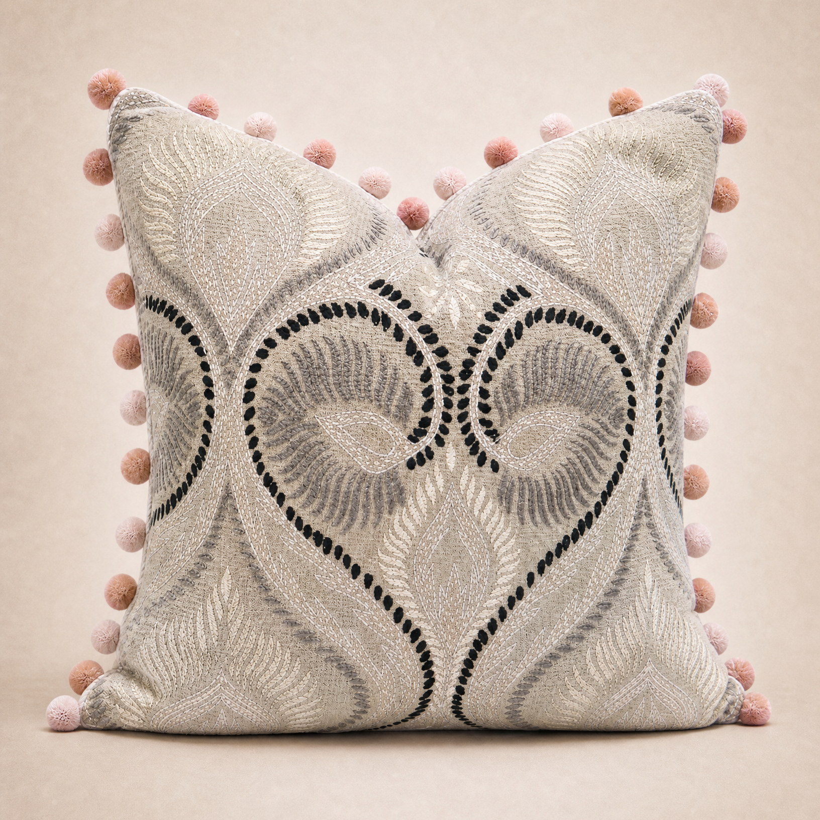 A square decorative throw pillow in a light beige fabric with a textured damask pattern, featuring black embroidered heart motifs and a border of pink pom-poms, set against a solid beige background.