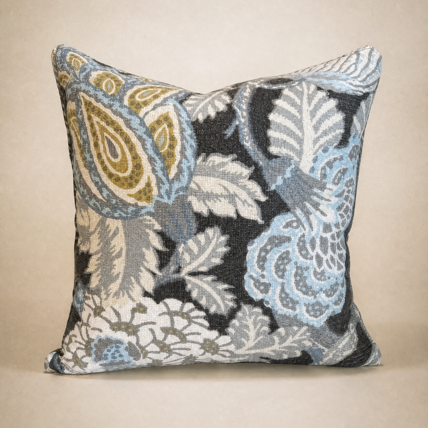 A square decorative throw pillow featuring an intricate woven floral and paisley pattern in light blue, beige, gray, and black, set against a solid beige background.