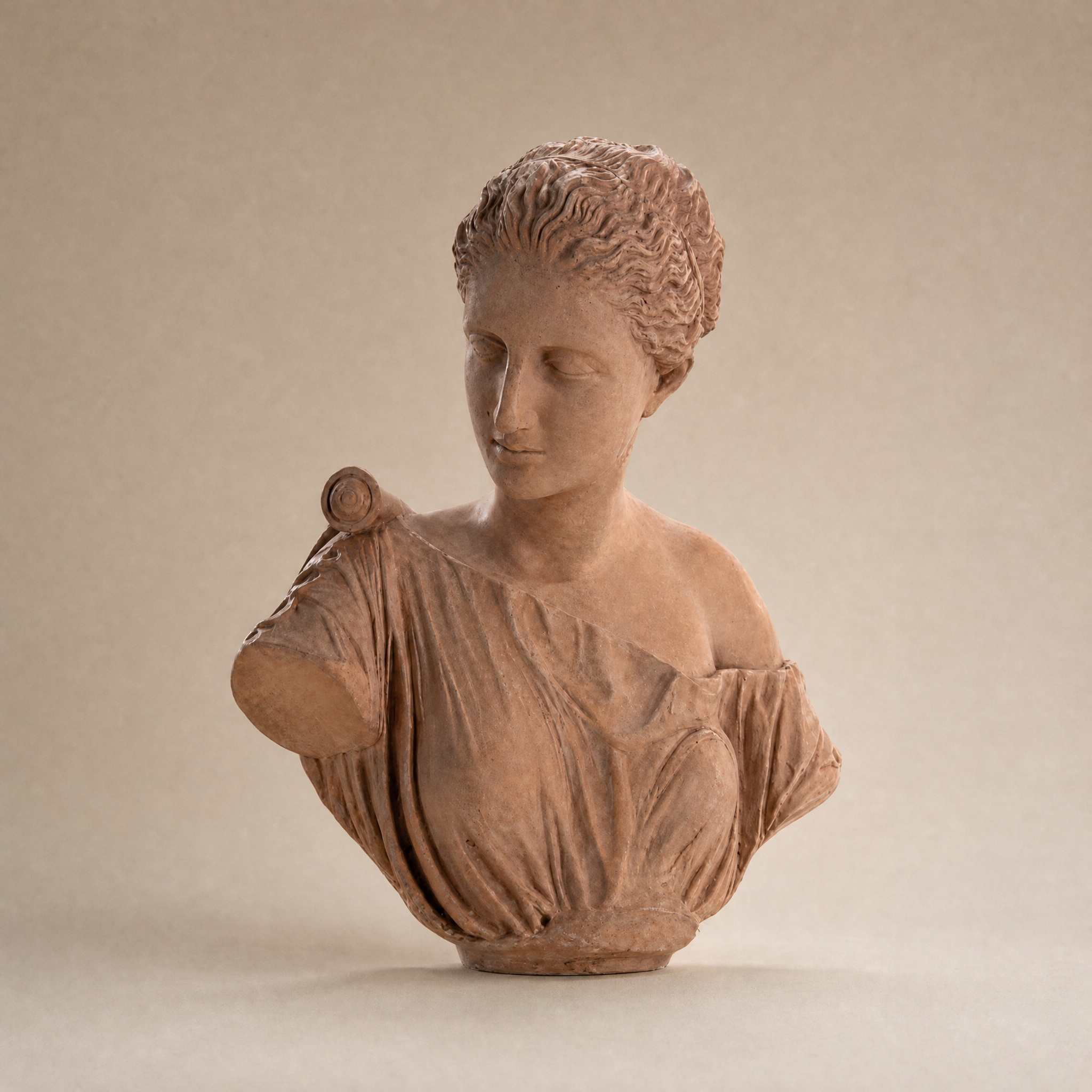 A terracotta-style classical female bust sculpture with detailed hair and draped fabric, photographed against a plain warm beige background.